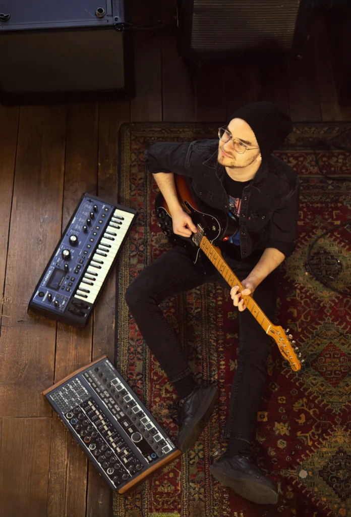 Mężczyzna w czarnej czapce siedzący na dywanie i grający na gitarze.